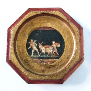 May include: An octagonal decorative tray with a painted scene inside a circular frame. The tray has a red border, a gold patterned inner border, and a gold interior. The painted scene depicts two figures with a deer.