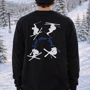 Op de afbeelding: Zwart shirt met lange mouwen met een witte afbeelding van skiërs en de tekst "Fuel your Soul". De persoon draagt een donkerblauwe muts. De achtergrond toont een besneeuwd berglandschap.