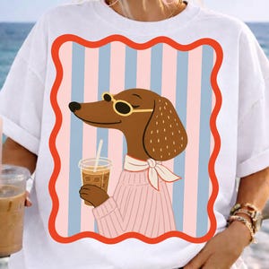 May include: White t-shirt with a cartoon dachshund wearing sunglasses and a pink sweater, holding an iced coffee. The dog is set against a striped background of pink and blue, framed by a red border. The shirt has short sleeves.