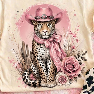 May include: Cream-colored t-shirt featuring a leopard wearing a pink cowboy hat and scarf, set against a pink backdrop with floral and cactus accents. The leopard has a detailed spotted coat. The design includes a large pink rose and other floral elements.