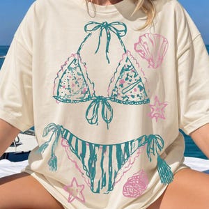 May include: Cream-colored t-shirt featuring a hand-drawn bikini design in teal and pink. The design includes a bikini top with floral patterns and a striped bottom, along with seashell and starfish accents. The text "Club Cheriee" is in the upper right corner.
