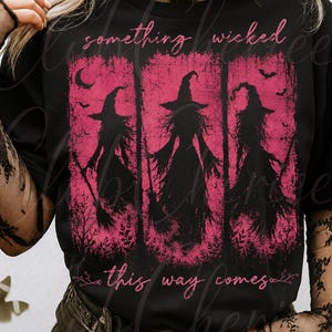 May include: A black t-shirt featuring a graphic print of three silhouettes of witches in pointed hats holding broomsticks. The design is set against a distressed pink background with bats and crescent moons, with the text something wicked this way comes.