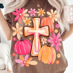 May include: A brown t-shirt featuring a central graphic of a cross surrounded by pink and orange pumpkins and various flowers in shades of pink, orange, and yellow. The design has a hand-painted aesthetic with vibrant colors and floral accents.
