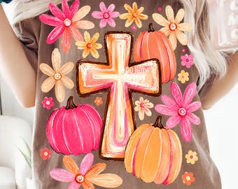 May include: A brown t-shirt featuring a central graphic of a cross surrounded by pink and orange pumpkins and various flowers in shades of pink, orange, and yellow. The design has a hand-painted aesthetic with vibrant colors and floral accents.