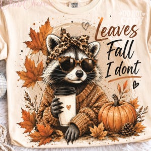 Puede incluir: Una camiseta color crema con el gráfico de un mapache que lleva una diadema de estampado de leopardo y gafas de sol, sosteniendo un vaso de café. El diseño incluye hojas de otoño naranjas, una calabaza y el texto Leaves Fall I dont.