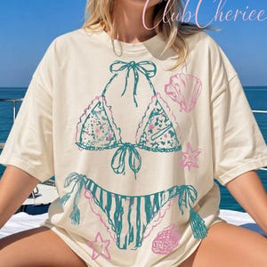 May include: Cream-colored t-shirt featuring a hand-drawn bikini design in teal and pink. The design includes a bikini top with floral patterns and a striped bottom, along with seashell and starfish accents. The text "Club Cheriee" is in the upper right corner.
