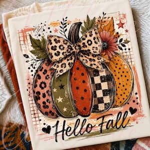 May include: A cream-colored t-shirt featuring a graphic of a pumpkin decorated with leopard print, polka dots, and checkered patterns. The pumpkin is topped with a large leopard print bow and autumn leaves. The text "Hello Fall" is printed below.