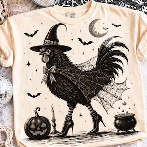 May include: A cream-colored t-shirt featuring a black graphic of a chicken wearing a witch hat, a polka-dot bow tie, a spiderweb cape, and high-heeled boots. The design includes a jack-o'-lantern, a candle, a cauldron, bats, and a crescent moon.