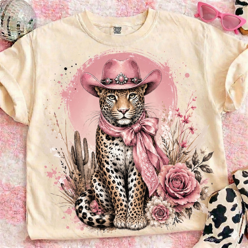 May include: Cream-colored t-shirt featuring a leopard wearing a pink cowboy hat and scarf, set against a pink backdrop with floral and cactus accents. The leopard has a detailed spotted coat. The design includes a large pink rose and other floral elements.