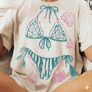 Puede incluir: Camiseta color crema con un dise&ntilde;o de bikini dibujado a mano en color turquesa y rosa. La parte superior del bikini tiene detalles florales y la parte inferior rayas verticales. Gr&aacute;ficos de conchas marinas y estrellas rosas dispersos.