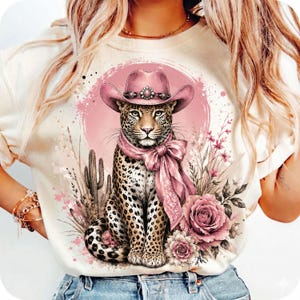 May include: A cream-colored t-shirt featuring a graphic print of a leopard wearing a pink cowboy hat and a matching pink neck scarf. The leopard is surrounded by pink watercolor splashes, a cactus, and pink roses on a circular pink background.