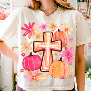 May include: A cream-colored short-sleeve t-shirt featuring a central graphic of a cross surrounded by pink and orange pumpkins and various colorful flowers in shades of pink, orange, and yellow. The design is printed on the front of the shirt.