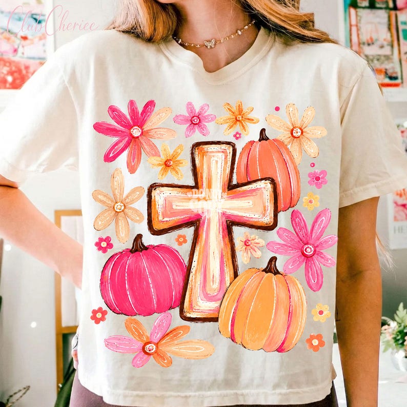 May include: A cream-colored short-sleeve t-shirt featuring a central graphic of a cross surrounded by pink and orange pumpkins and various colorful flowers in shades of pink, orange, and yellow. The design is printed on the front of the shirt.