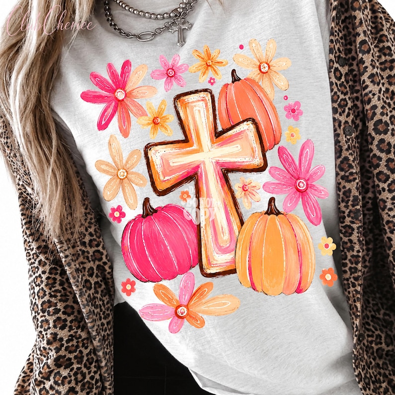 May include: A light gray t-shirt featuring a graphic print of a central cross surrounded by orange and pink pumpkins and various colorful flowers in shades of pink, orange, and yellow. The design is layered over a leopard print button-down shirt.