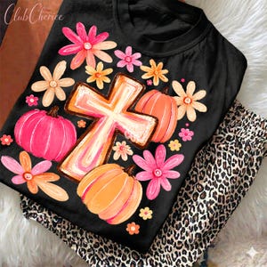 May include: A black t-shirt featuring a central graphic of a cross surrounded by pink and orange pumpkins and various flowers in shades of pink, orange, and yellow. The design is printed on the front of the shirt, which has a crew neckline.