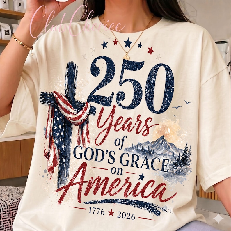 K&ouml;nnte beinhalten: Ein cremefarbenes T-Shirt mit einer Grafik, die ein mit einer amerikanischen Flagge drapiertes Kreuz, eine Berglandschaft und den Text 250 Years of GOD'S GRACE on America 1776-2026 zeigt. Das Design enth&auml;lt blaue und rote Schriftz&uuml;ge mit Sternen.