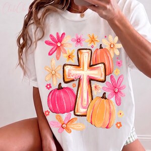 May include: A white short-sleeve t-shirt featuring a central graphic of a cross surrounded by orange and pink pumpkins and various colorful flowers in shades of yellow, pink, and orange. The design has a hand-drawn, artistic aesthetic.