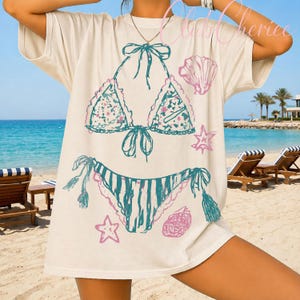 Puede incluir: Camiseta color crema con un dise&ntilde;o de bikini turquesa y rosa, adornada con conchas marinas y estrellas de mar. El dise&ntilde;o del bikini incluye patrones florales y detalles de borlas, evocando un ambiente playero y veraniego. La camiseta tiene mangas cortas.