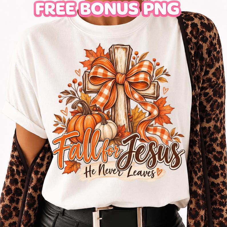 May include: A white t-shirt featuring a graphic of a wooden cross adorned with an orange and white checkered bow, surrounded by autumn leaves and pumpkins. The text reads Fall for Jesus He Never Leaves. A leopard print cardigan is worn over the shirt.