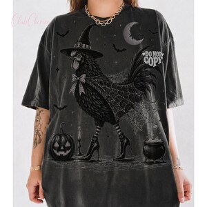 May include: A charcoal gray oversized t-shirt featuring a graphic of a chicken wearing a witch hat, a bow, and high-heeled boots. The design includes a spiderweb cape, a jack-o'-lantern, a cauldron, bats, and a crescent moon. The text DO NOT COPY is visible.