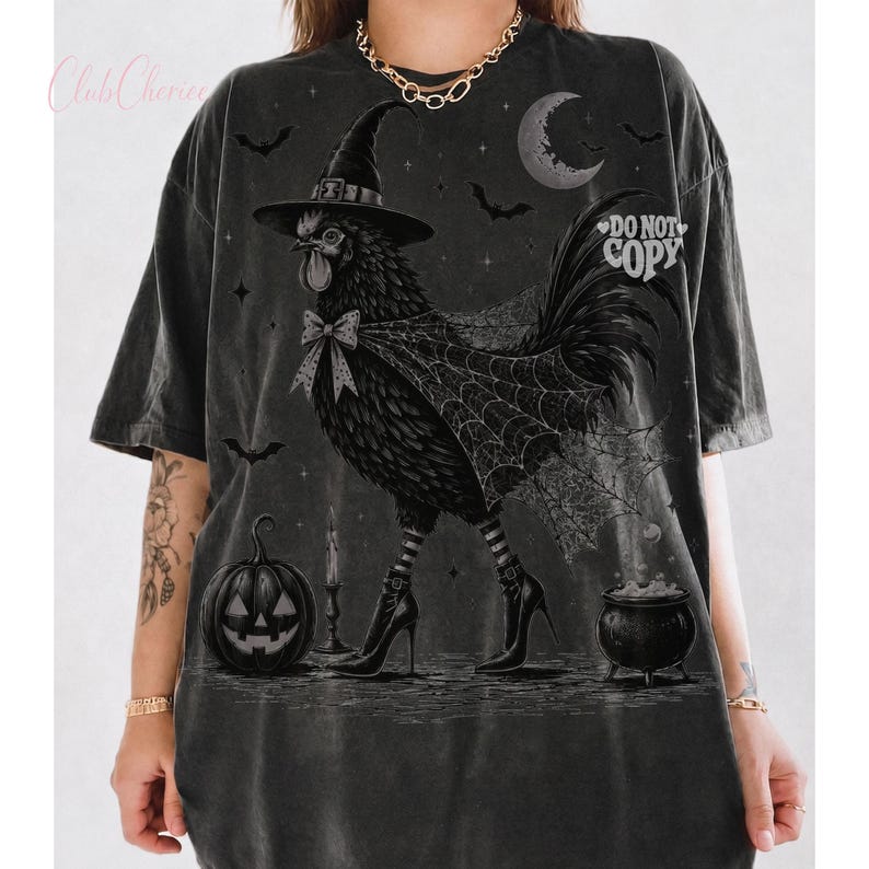 May include: A charcoal gray oversized t-shirt featuring a graphic of a chicken wearing a witch hat, a bow, and high-heeled boots. The design includes a spiderweb cape, a jack-o'-lantern, a cauldron, bats, and a crescent moon. The text DO NOT COPY is visible.