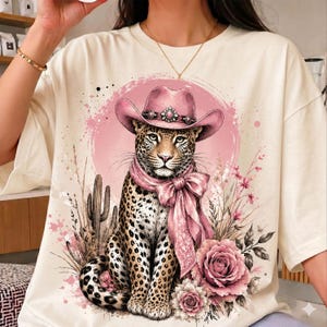 May include: A cream-colored t-shirt featuring a graphic print of a leopard wearing a pink cowboy hat and a matching pink neck scarf. The leopard is surrounded by pink floral accents, desert cacti, and a circular pink watercolor-style background.