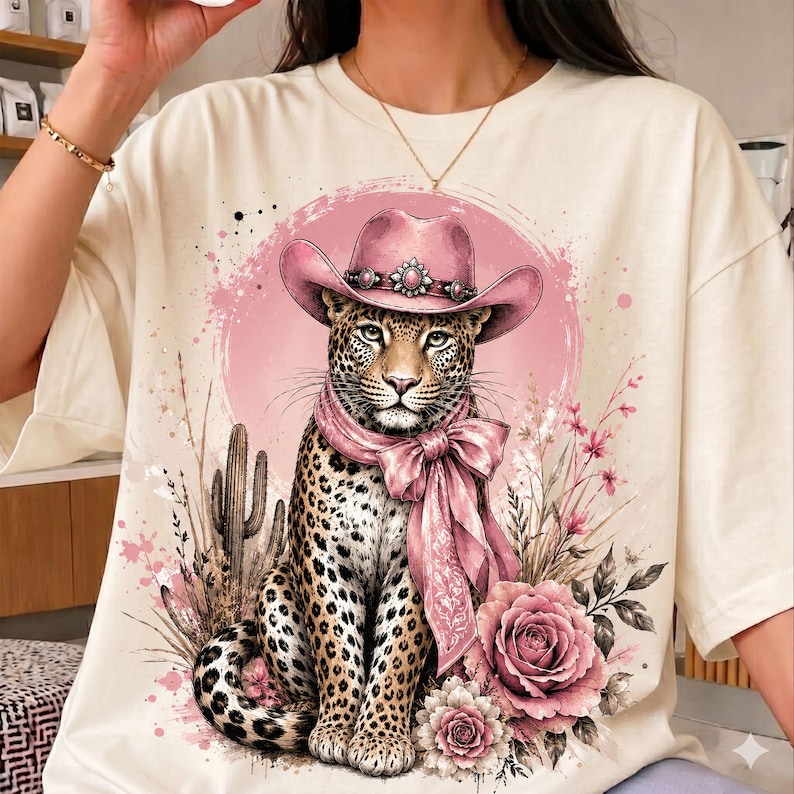 May include: A cream-colored t-shirt featuring a graphic print of a leopard wearing a pink cowboy hat and a matching pink neck scarf. The leopard is surrounded by pink floral accents, desert cacti, and a circular pink watercolor-style background.