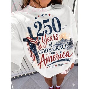 K&ouml;nnte beinhalten: Ein wei&szlig;es Sweatshirt mit Rundhalsausschnitt und einem Grafikdruck, der ein Kreuz mit einer amerikanischen Flagge, eine Berglandschaft und den Text 250 Years of GOD'S GRACE on America 1776 2026 zeigt. Das Design nutzt blaue, rote und schwarze Schrift.