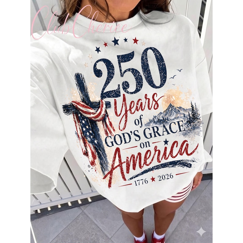 K&ouml;nnte beinhalten: Ein wei&szlig;es Sweatshirt mit Rundhalsausschnitt und einem Grafikdruck, der ein Kreuz mit einer amerikanischen Flagge, eine Berglandschaft und den Text 250 Years of GOD'S GRACE on America 1776 2026 zeigt. Das Design nutzt blaue, rote und schwarze Schrift.