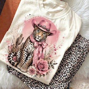 May include: A cream-colored t-shirt featuring a graphic print of a leopard wearing a pink cowboy hat and a matching pink neck scarf. The leopard is surrounded by pink roses, desert cacti, and a circular pink background with paint splatters.