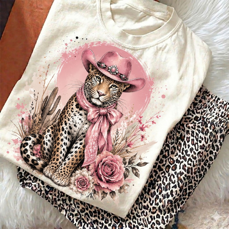 May include: A cream-colored t-shirt featuring a graphic print of a leopard wearing a pink cowboy hat and a matching pink neck scarf. The leopard is surrounded by pink roses, desert cacti, and a circular pink background with paint splatters.