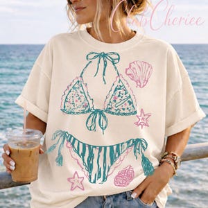 Puede incluir: Camiseta color crema con un dise&ntilde;o de bikini turquesa y rosa con acentos de conchas marinas y estrellas de mar. La camiseta gr&aacute;fica tiene un corte holgado y mangas cortas, perfecta para un tema de playa o verano.