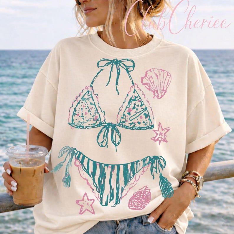 Puede incluir: Camiseta color crema con un dise&ntilde;o de bikini turquesa y rosa con acentos de conchas marinas y estrellas de mar. La camiseta gr&aacute;fica tiene un corte holgado y mangas cortas, perfecta para un tema de playa o verano.