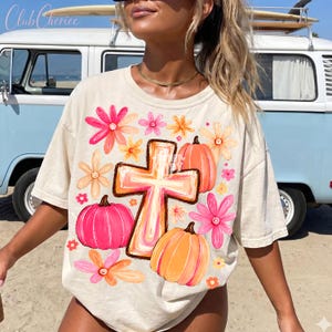 May include: A cream-colored t-shirt featuring a central graphic of a cross surrounded by pink and orange pumpkins and various colorful flowers in shades of pink, orange, and yellow. The design is printed on the front of the short-sleeved garment.