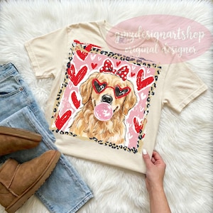 May include: Cream-colored t-shirt featuring a graphic of a golden retriever wearing heart-shaped sunglasses and a bow, blowing a bubblegum bubble. The design includes red hearts and a leopard print border.