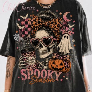 May include: A black t-shirt featuring a graphic of a skeleton wearing heart-shaped sunglasses and a leopard print bow. The design includes a ghost, a pumpkin, a coffee cup with the text Spooky But Cute, and the text Spooky Season.