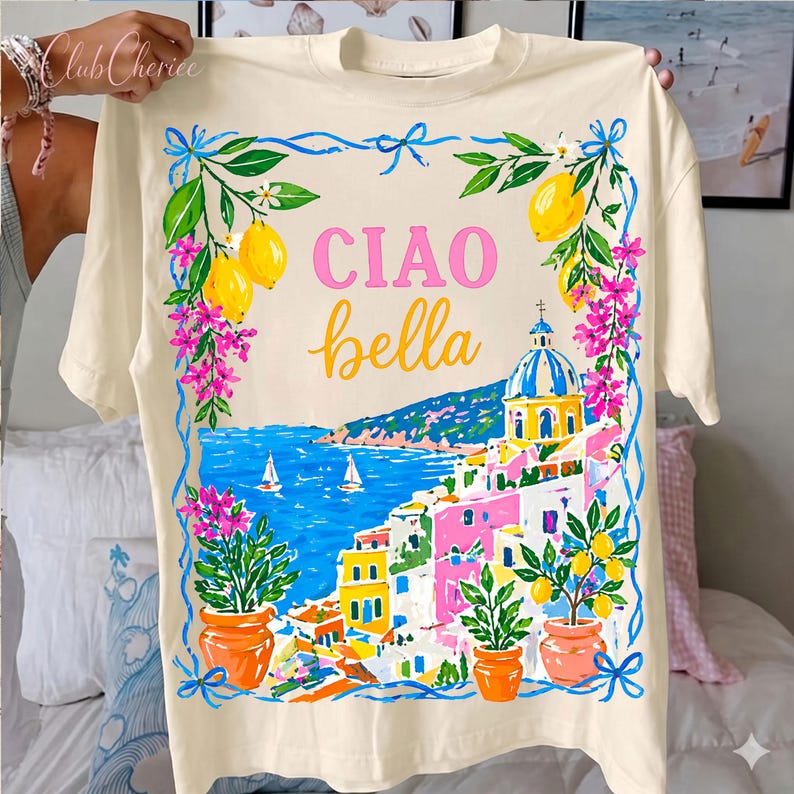 Może przedstawiać: Kremowy t-shirt z kolorowym włoskim motywem nadmorskim, z napisami "CIAO bella" w kolorze r&oacute;żowym i ż&oacute;łtym. Projekt zawiera cytryny, kwiaty i niebieskie morze z żagl&oacute;wkami. Koszulka ma okrągły dekolt i kr&oacute;tkie rękawy.