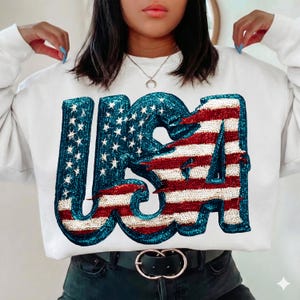 May include: White sweatshirt with the word "USA" in a sequined design. The letters are filled with the American flag's stars and stripes in red, white, and blue. The sweatshirt has a crew neck and long sleeves.