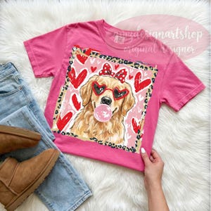 May include: Pink t-shirt featuring a graphic of a golden retriever wearing heart-shaped sunglasses and a bow, blowing a bubblegum bubble. The design includes red hearts and a leopard print border. The shirt is paired with blue jeans and brown boots.