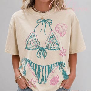 Puede incluir: Camiseta beige con un dise&ntilde;o de bikini turquesa y rosa. La parte superior del bikini tiene un estampado floral y ribete de encaje, con la parte inferior a juego. Tambi&eacute;n se imprimen gr&aacute;ficos de conchas marinas y estrellas de mar rosas en la camiseta.