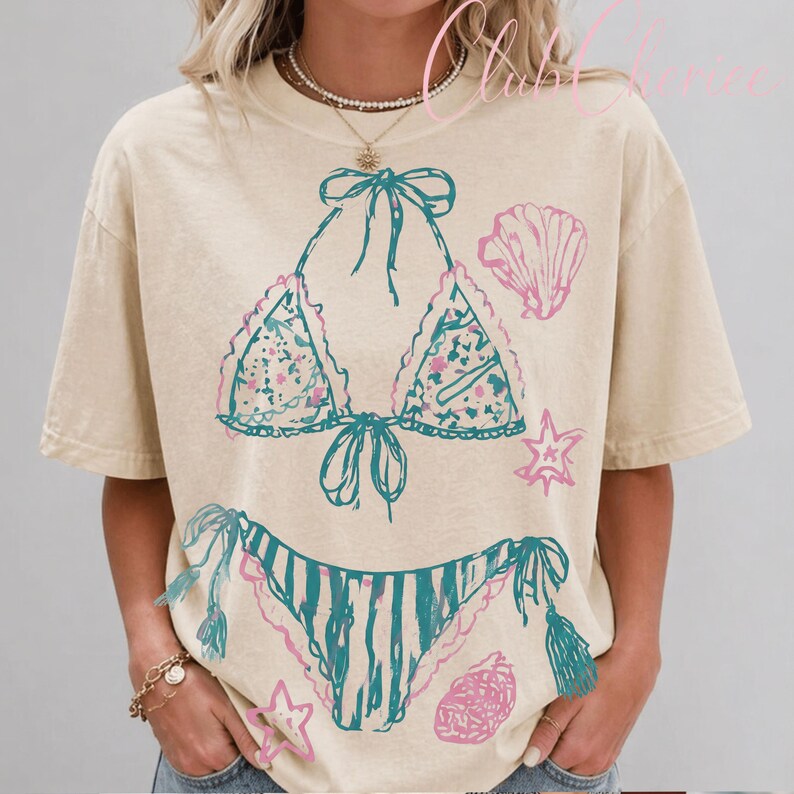 Puede incluir: Camiseta beige con un dise&ntilde;o de bikini turquesa y rosa. La parte superior del bikini tiene un estampado floral y ribete de encaje, con la parte inferior a juego. Tambi&eacute;n se imprimen gr&aacute;ficos de conchas marinas y estrellas de mar rosas en la camiseta.