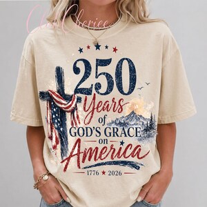 K&ouml;nnte beinhalten: Ein beiges Kurzarm-T-Shirt mit einem Grafikdruck, der ein Kreuz mit einer amerikanischen Flagge, eine Berglandschaft und den Text 250 Years of GOD'S GRACE on America 1776 2026 zeigt. Das Design verwendet marineblaue, rote und wei&szlig;e Farben.