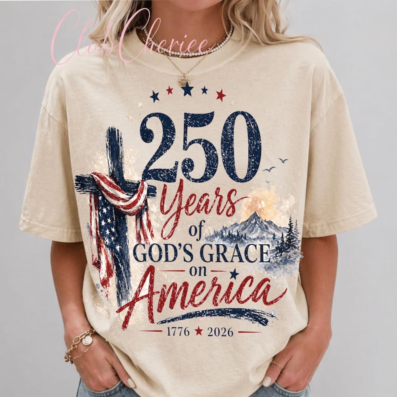 K&ouml;nnte beinhalten: Ein beiges Kurzarm-T-Shirt mit einem Grafikdruck, der ein Kreuz mit einer amerikanischen Flagge, eine Berglandschaft und den Text 250 Years of GOD'S GRACE on America 1776 2026 zeigt. Das Design verwendet marineblaue, rote und wei&szlig;e Farben.
