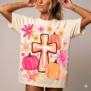 May include: A cream-colored short-sleeve t-shirt featuring a central graphic of a cross surrounded by pink and orange pumpkins and various colorful flowers in shades of pink, orange, and yellow. The text Club Cheriee is printed in the top left corner.