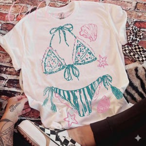 Puede incluir: Camiseta blanca con un dise&ntilde;o de bikini turquesa y rosa. La parte superior e inferior del bikini est&aacute;n ilustradas con patrones florales y de rayas, junto con acentos de conchas marinas y estrellas. La camiseta es de corte casual.
