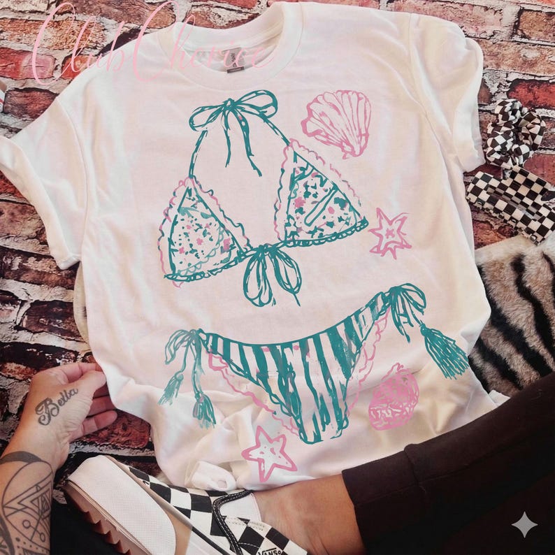 Puede incluir: Camiseta blanca con un dise&ntilde;o de bikini turquesa y rosa. La parte superior e inferior del bikini est&aacute;n ilustradas con patrones florales y de rayas, junto con acentos de conchas marinas y estrellas. La camiseta es de corte casual.