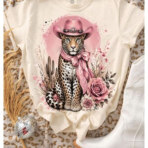 May include: A cream-colored t-shirt featuring a graphic print of a leopard wearing a pink cowboy hat and a matching pink neck scarf. The leopard is surrounded by pink floral elements, desert cacti, and a circular pink background design.