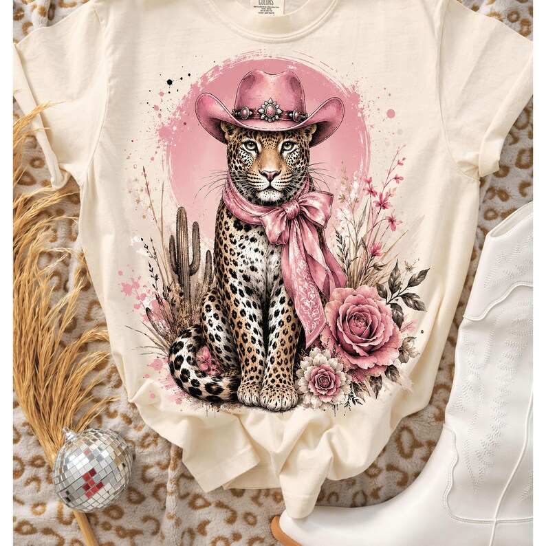 May include: A cream-colored t-shirt featuring a graphic print of a leopard wearing a pink cowboy hat and a matching pink neck scarf. The leopard is surrounded by pink floral elements, desert cacti, and a circular pink background design.