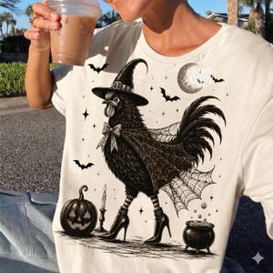 May include: A cream-colored t-shirt featuring a graphic of a black rooster dressed as a witch, wearing a pointed hat, a bow tie, a spiderweb cape, and high-heeled boots. The design includes a jack-o'-lantern, a candle, a cauldron, and bats.
