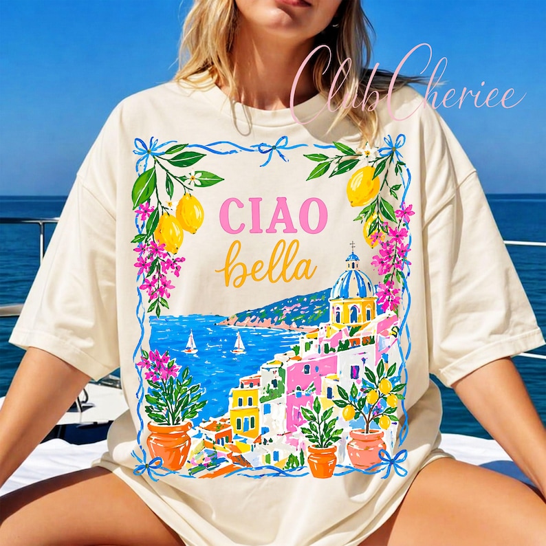May include: Cream-colored t-shirt featuring a colorful Italian coastal scene with the words "CIAO bella" in pink and yellow. The design includes lemons, flowers, and a blue sea with boats. The shirt has a relaxed fit.